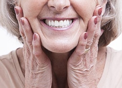 An elderly woman smiling, showing her bright white dentures with her hands on her cheeks